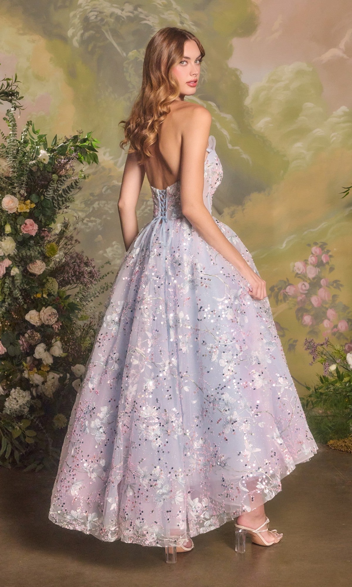 A woman in the Andrea & Leo A1485 Strapless Sequin Prom Dress stands with her back to the camera, surrounded by lush greenery and flowers, her dress evoking the elegance of an enchanting garden scene.